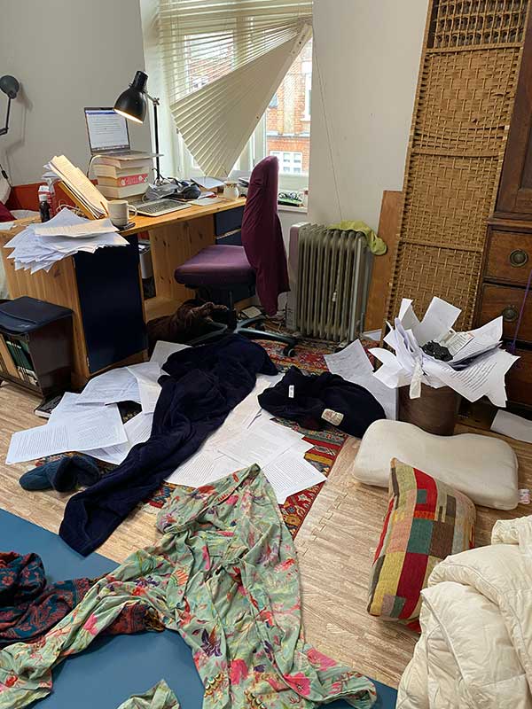 writing-opt A photo showing where Emily Woof writes; a laptop balanced on a pile of books. There is paper and clothes strewn across the floor.